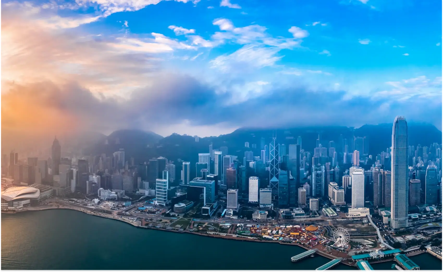 A panoramic view of the Hong Kong skyline with clouds and sunlight illuminating the cityscape